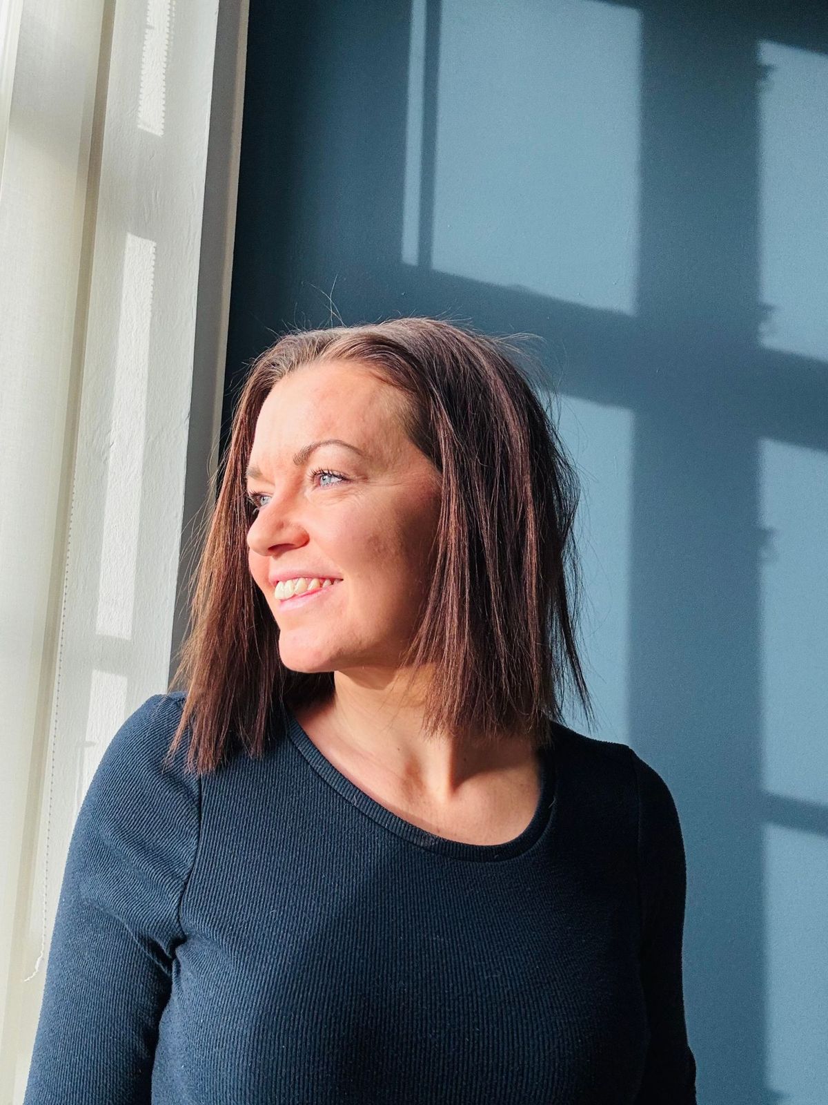 Portræt af Alice Heilesen, der smiler og kigger ud ad vinduet foran en blå væg